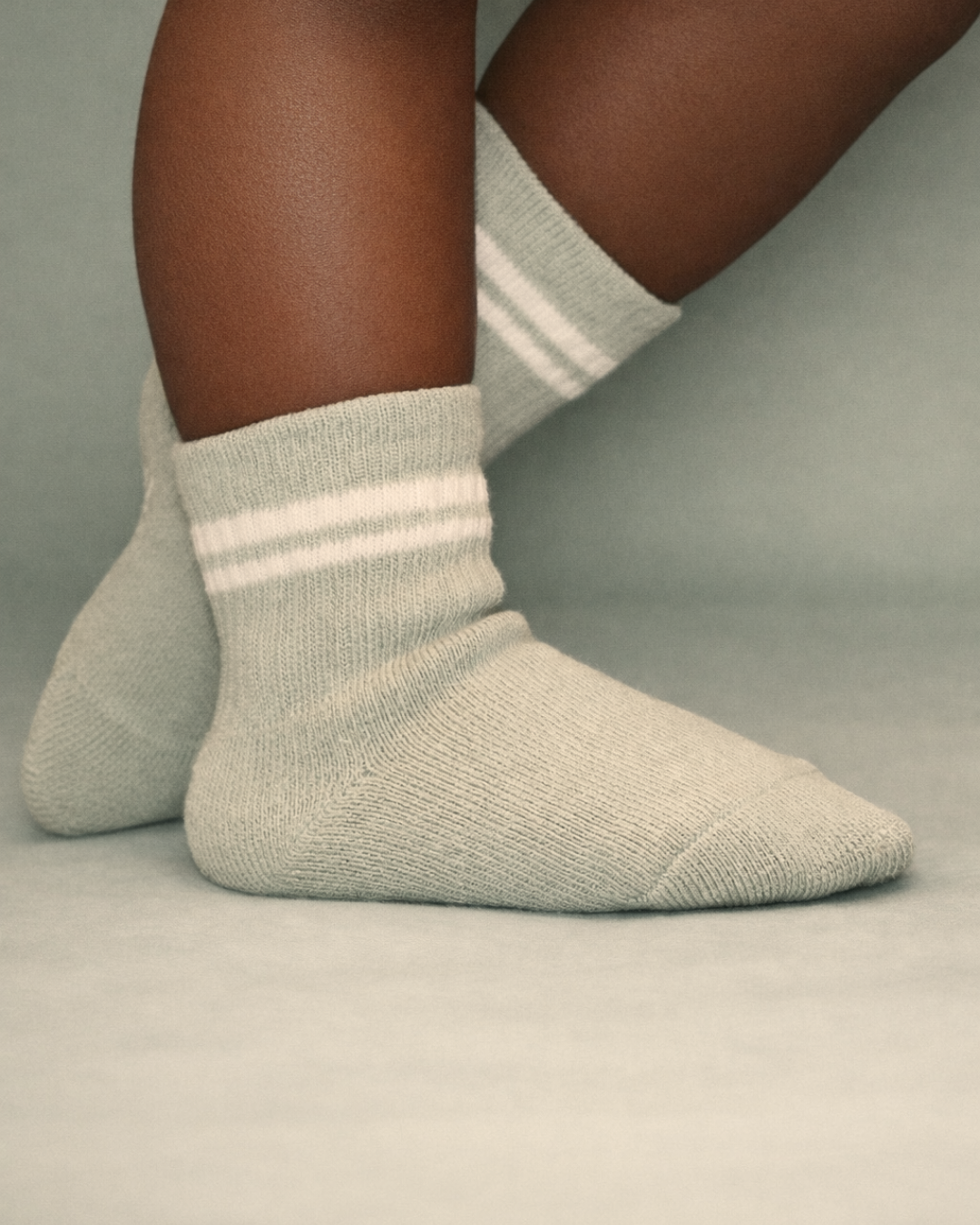 Baby with a dark skin tone wearing a pair of sage baby crew socks with silicone grips on a sage background