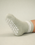 Baby with a medium skin tone wearing a sage green baby crew sock with white heart and dot silicone grips on a sage background