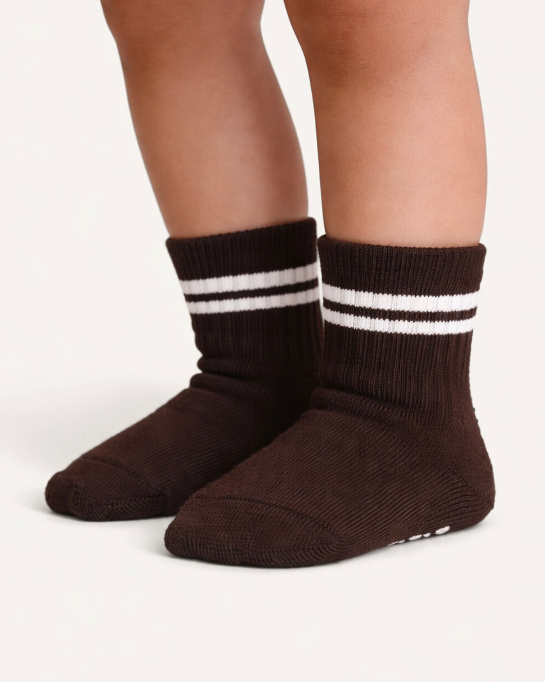 Pair of dark brown baby crew socks with white stripes and silicone grips on an off-white background